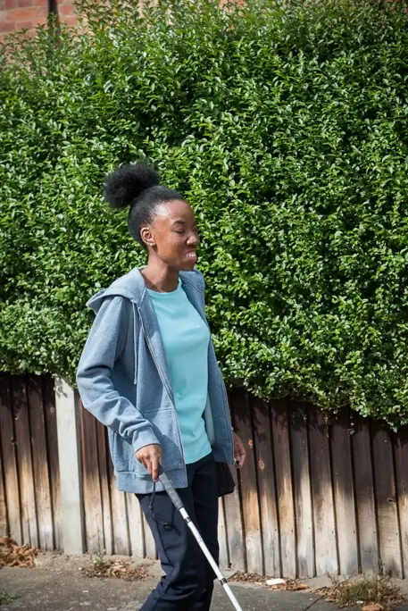Blind psychotherapeutic counsellor, Sandra, walking along wooden fence with her white cane wearing a blue t-shirt and a grey zip-up hoodie